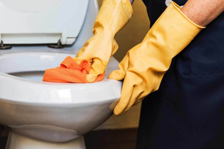 a person cleaning toilet seat wearing yellow gloves and cleaning toilet seat with orange color cloth.