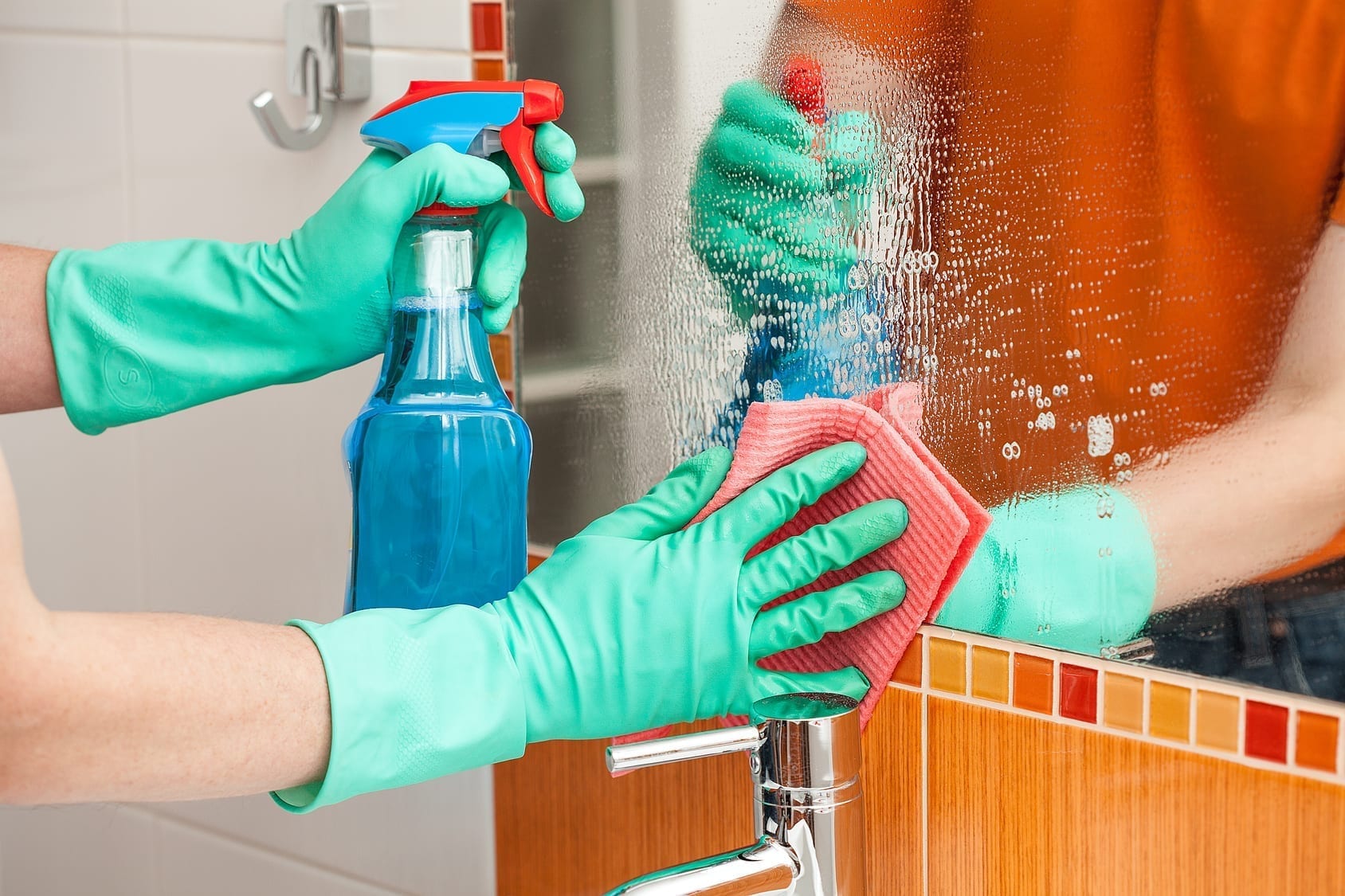 close shot of hands wearing green color protective gloves holding a ClO2 Spray bottle and a sponge cleaning and sanitizing mirror.