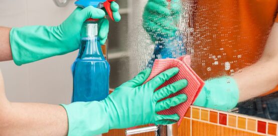 close shot of hands wearing green color protective gloves holding a ClO2 Spray bottle and a sponge cleaning and sanitizing mirror.