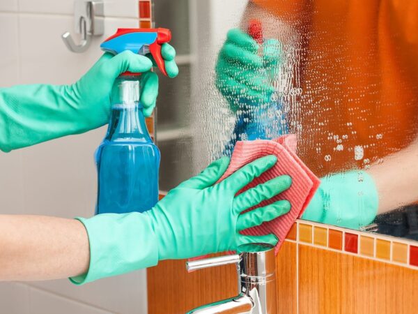 close shot of hands wearing green color protective gloves holding a ClO2 Spray bottle and a sponge cleaning and sanitizing mirror.