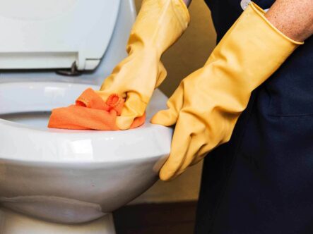 a person cleaning toilet seat wearing yellow gloves and cleaning toilet seat with orange color cloth.